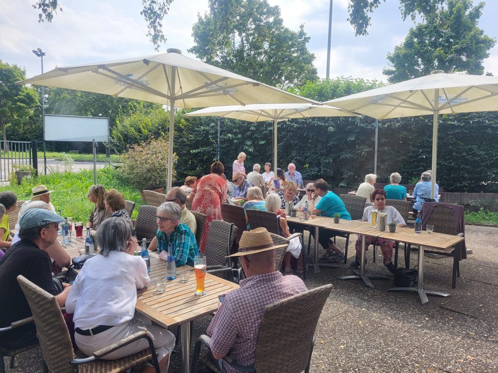Menschen sitzen unter Sonnenschirmen an Tischen beim Sommerfest in Karlsruhe, genießen Getränke und Unterhaltung im Freien