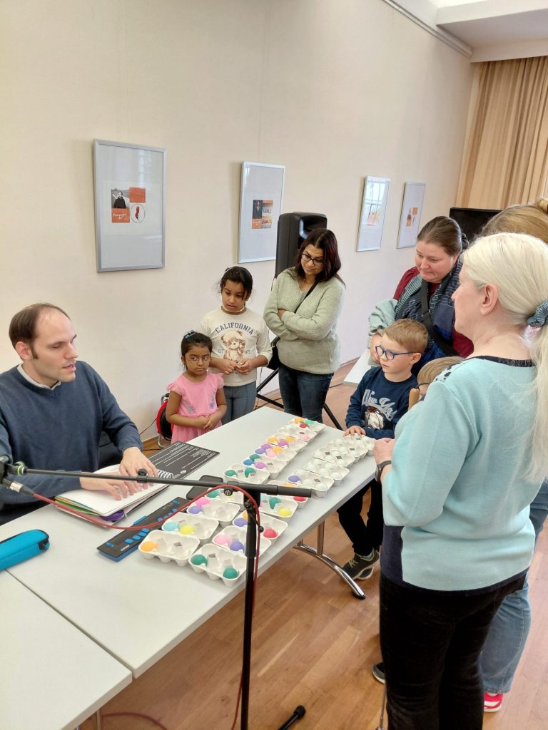 Mehrere Personen, darunter einige Kinder und Erwachsene, stehen um einen Tisch. Die Personen blicken auf den Tisch, auf dem auch ein Mikrofonständer zu sehen ist. Vor dem Tisch sitzt ein Mann, der mit seinen Händen ein Kinderbuch für blinde- und sehbehinderte Kinder ertastet und daraus vorliest. Im Hintergrund hängen drei gerahmte Bilder an der Wand, und rechts ist ein Fenster mit hellen Vorhängen zu erkennen.