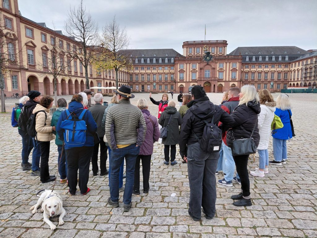 Eine Gruppe von Menschen steht auf einem gepflasterten Platz vor dem Barockschloss in Mannheim. Die Personen tragen überwiegend Jacken und Rucksäcke, einige haben Hüte oder Mützen. Eine Person in der Mitte hebt eine Hand, sie spricht und zeigt etwas. Vor der Gruppe liegt ein weißer Hund auf dem Boden. Im Hintergrund ist das große, mehrstöckige Barockschloss mit zahlreichen Fenstern und einem Uhrenturm zu sehen. Der Himmel ist bewölkt, und einige Bäume mit gelben Blättern stehen am Rand des Platzes.