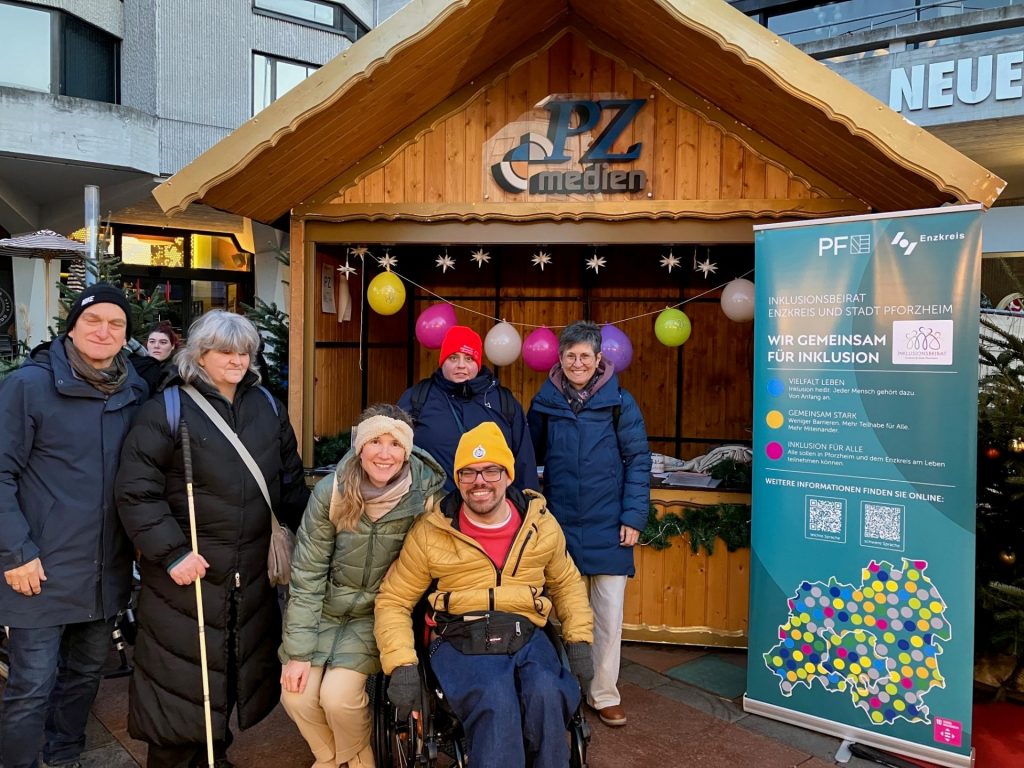 Vor einem kleinen, hölzernen Weihnachtsmarktstand mit dem Schild 'PZ medien' steht eine Gruppe von sechs Personen, die winterliche Kleidung tragen. Rechts neben der Gruppe befindet sich ein aufrechter, türkisfarbener Informationsaufsteller mit dem Text 'WIR GEMEINSAM FÜR INKLUSION'. Der Stand ist mit bunten Luftballons und kleinen weißen Sternen dekoriert.