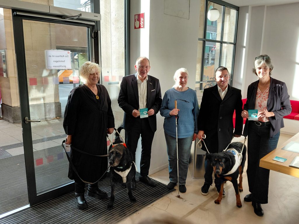 Fünf Personen stehen in einem hellen Innenraum vor großen Fenstern und einer Glastür. Zwei der Personen halten jeweils einen Blindenführhund an der Leine. Eine Person hält einen weißen Blindenstock.