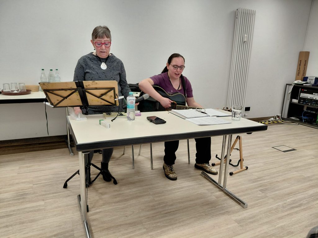 Zwei Personen sitzen an einem hellen Tisch mit Notenständer, Gitarre und Getränkeflaschen im Hintergrund.