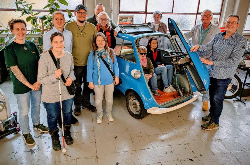 Mehrere Personen stehen und sitzen um ein kleines, blaues Auto mit geöffneter Tür in einem Innenraum.