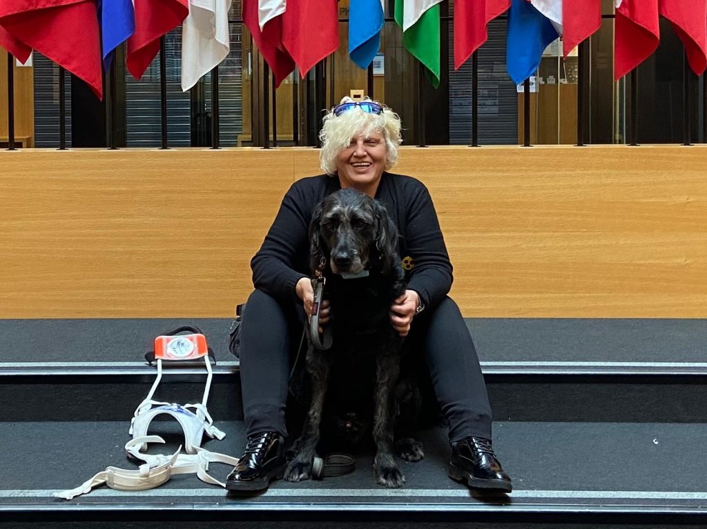 Die Leiterin der Führundhaltergrppe sitzt auf einer Treppe und hält einen großen, dunkelhaarigen Hund vor sich an der Leine. Sie trägt dunkle Kleidung und glänzende schwarze Schuhe. Neben ihr liegt ein weißes Führgeschirr für Blindenführhunde. Im Hintergrund hängen zahlreiche bunte Flaggen von der Decke.