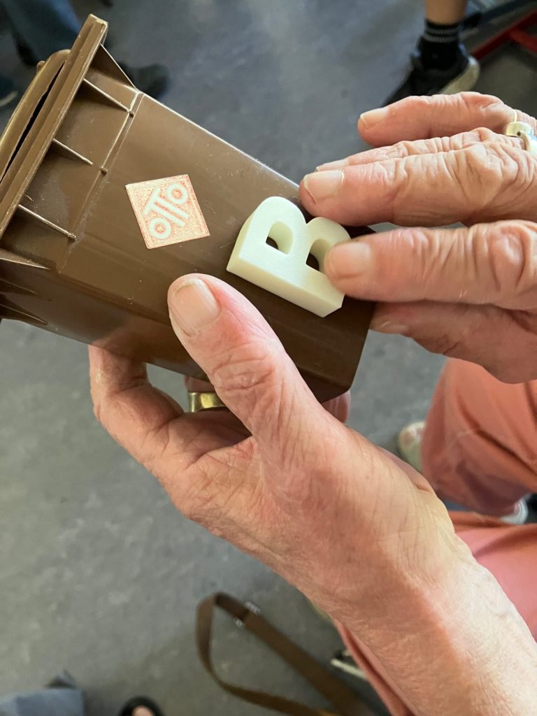 Eine Person hält eine kleine braune Modell-Mülltonne in der Hand. Mit den Fingern wird ein weißer, dreidimensionaler Buchstabe B ertastet, der auf der Vorderseite der Tonne angebracht ist.